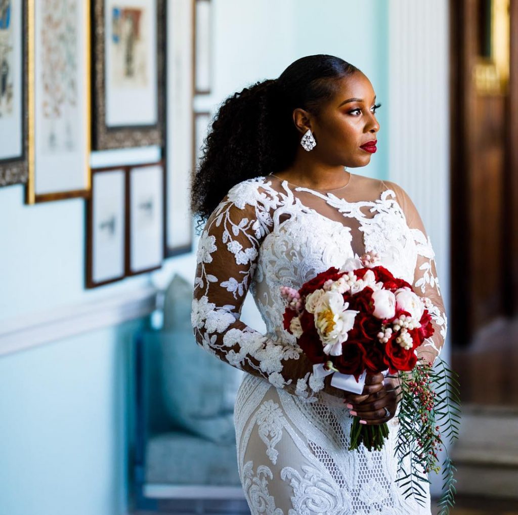 Black woman dressed in a white wedding dress holding red & white flowers with wall frames and furniture in the background